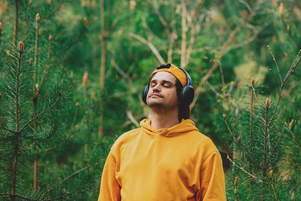 Homme élégant en sweat à capuche écouter de la musique dans les écouteurs dans la forêt de pins - Photo, image