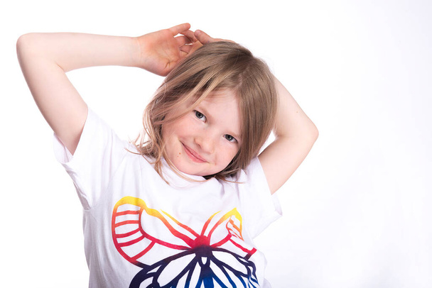 a little smiling girl in a white t-shirt holds her hands behind her head and looks at the camera  - Photo, image