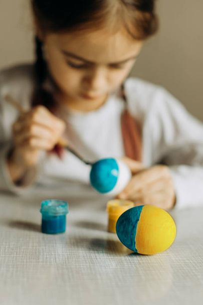 Portrait of a Ukrainian girl. the child paints the egg blue and yellow. The child loves and supports his country. Cute little child girl painting with blue and yellow colors Easter eggs. Hands of a girl with a easter egg. Close-up. - Фото, зображення