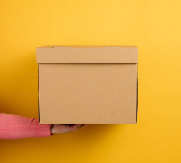 mano femenina sosteniendo cerrada caja de cartón marrón sobre fondo amarillo, entrega - Foto, Imagen