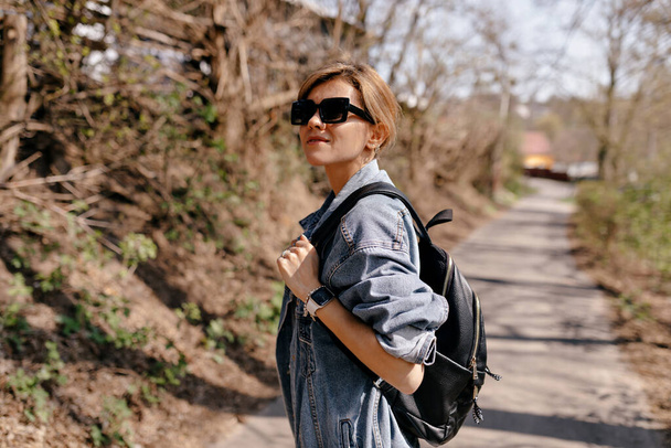 Pretty charming girl with light hair in sunglasses and denim jacket with backpack is walking on the forest trail in warm spring day. High quality photo - Foto, Imagen