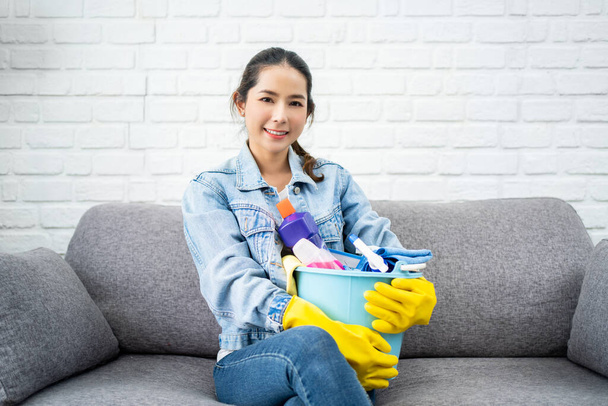Portrait of beautiful Asian woman female in yellow gloves holding bucket with cleaning sprays and tools sitting on sofa after cleaning home. Domestic work housekeeping concept. - Photo, image