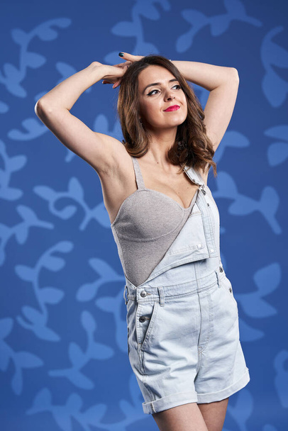 Happy young woman in denim overalls, studio shot over blue floral background - Foto, immagini