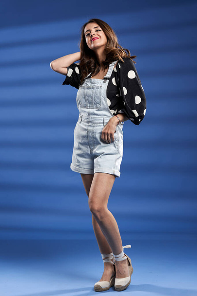 Beautiful young woman in denim overalls against blue background, studio shot - Foto, afbeelding