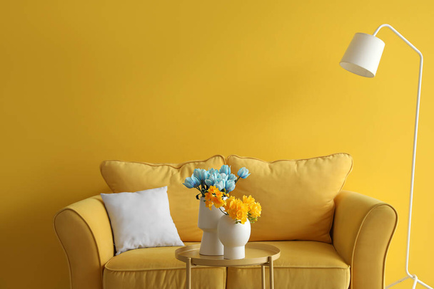 Утішна дивана і гарні квіти в кольорах українського прапора на столі біля кольорової стіни у вітальні. - Фото, зображення
