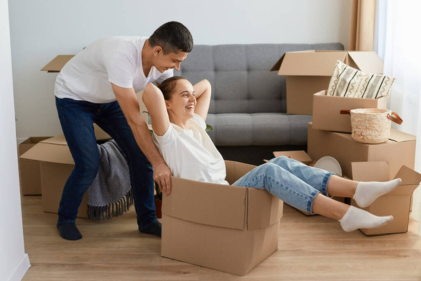 Portrait of positive delighted couple having fun laughing, relocating into new apartment, woman riding in cardboard box while man pushing it, family unpacking personal piles together. - Foto, imagen