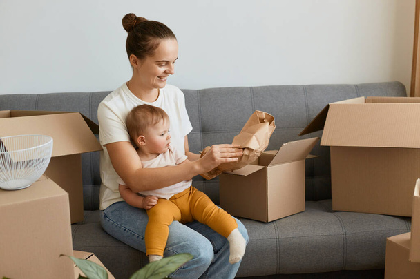 Image of smiling woman in white t shirt sitting on sofa surrounded with carton boxes with her personal piles and holding her infant baby, unpacking box with glassware. - Photo, Image