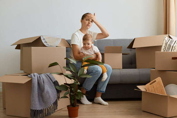 Indoor shot of tired exhausted dark haired woman weairng casual attire sitting on cough with her infant toddler daughter, unpacking personal piles in cardboard boxes. - Foto, imagen
