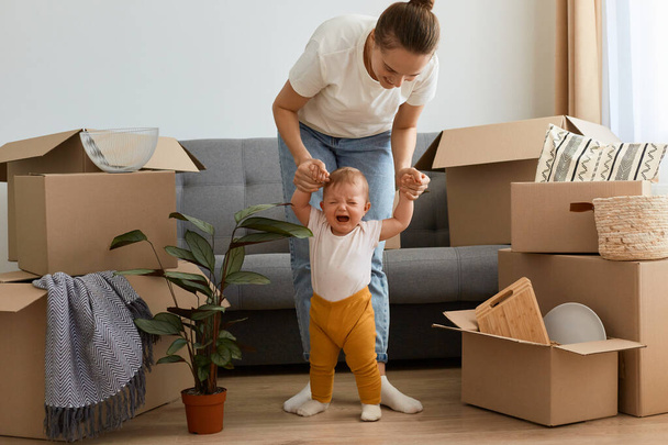 Indoor shot of dark haired attractive woman, young mother wearing white t shirt and jeans playing with her little baby daughter while relocating to a new house. - Foto, Imagen