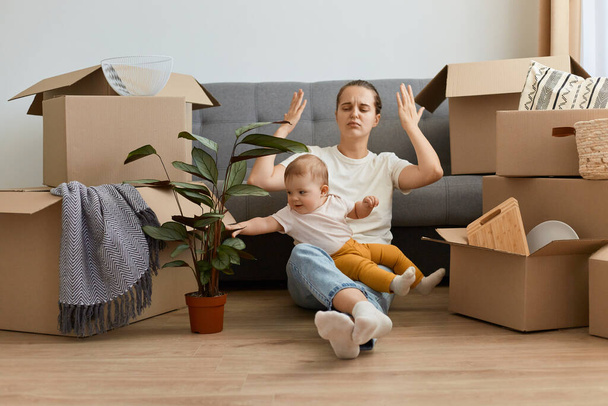 Portrait of Caucasian woman wearing white t shirt and jeans sitting on floor near sofa with her infant daughter, relocating to a new flat, being tired unpacking personal belongings with her baby. - Photo, image