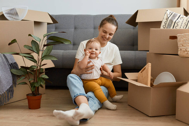 Full length portrait of satisfied female with dark hair wearing white t shirt playing with her daughter, sitting on floor surrounded with cardboard boxes, relocating. - Фото, изображение