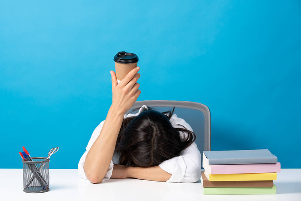 Woman sleeping head on desk showing disposable cup of coffee isolated on blue background. Caffeine addiction concept - Foto, imagen