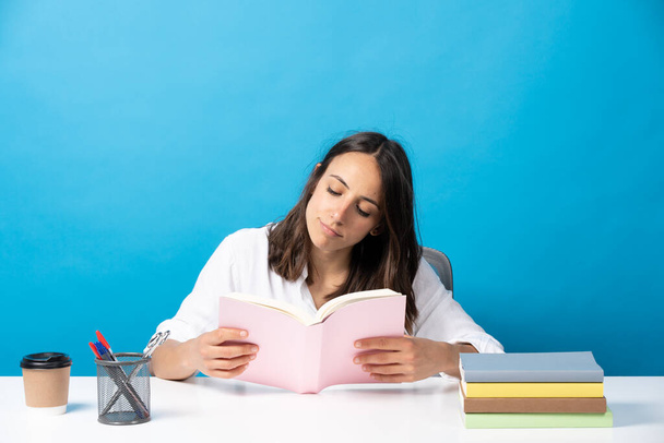 Μια νεαρή όμορφη Ισπανίδα διαβάζει ένα βιβλίο πίσω από ένα γραφείο απομονωμένο σε μπλε φόντο.. - Φωτογραφία, εικόνα