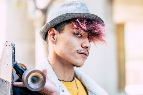 Portré alternatív megjelenés spanyol fiatal trendi fiú színes haj és kalap. Modern stílus divat tizenéves felnőtt nézi a kamera és a gazdaság gördeszka fórumon - Fotó, kép