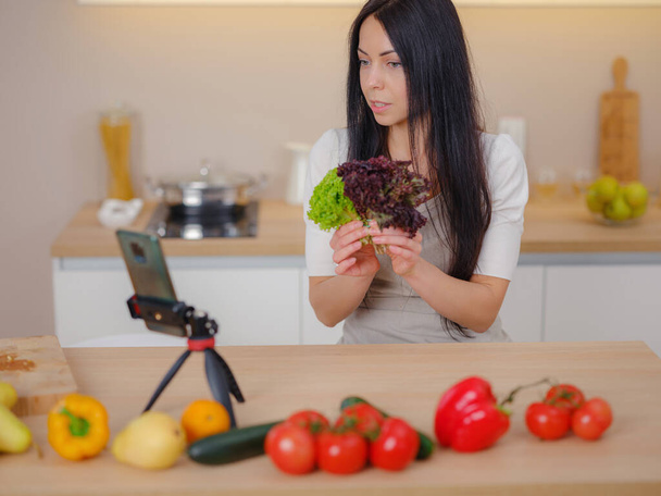 Красивая счастливая молодая женщина ведет блог на своем кухонном канале о здоровом образе жизни на кухне своего дома и смотрит на смартфон на штативе - Фото, изображение