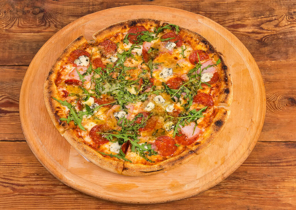 Pizza with salami and ham on the big round wooden pizza board on the old rustic wooden table - Photo, image