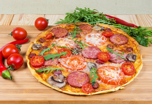 Pieczona pizza okrągła z salami, inne kiełbasy, pomidory i grzyby wśród świeżych warzyw na drewnianej desce do krojenia - Zdjęcie, obraz