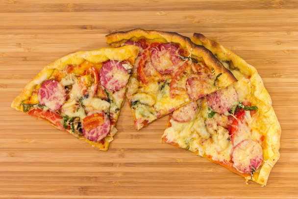 Kawałki pieczonej pizzy z salami, pomidorami i grzybami na drewnianej desce do krojenia, widok z góry - Zdjęcie, obraz