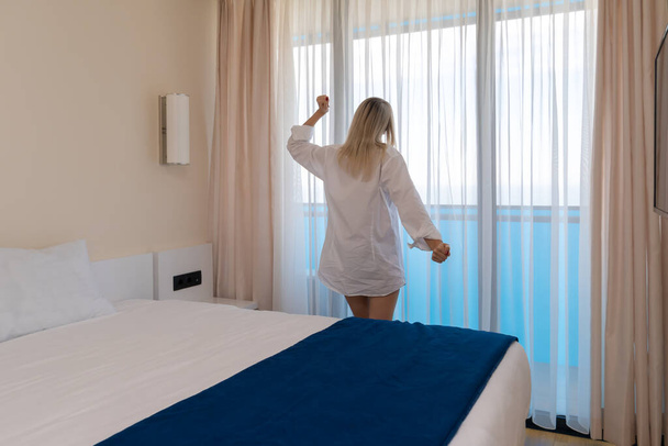 Jovem mulher loira delgada em uma camisa branca em um corpo nu fica na janela e se estende após uma boa noite de sono - Foto, Imagem