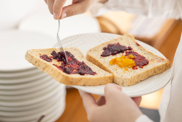 hand van toepassing zoete bessen fruit jam op een gesneden volkoren brood voor het ontbijt high energy maaltijd in de ochtend - Foto, afbeelding