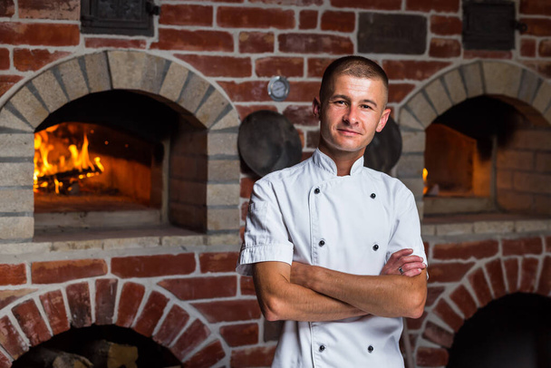 Portrait of a smiling pizza chef standing on the background of a wood-burning oven in the kitchen - 写真・画像