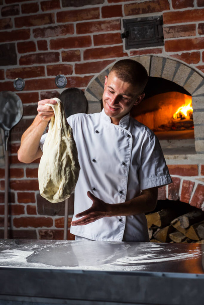 Chief chef kneads the dough on table with flour for cooking Italian pizza near a wood-burning oven in the kitchen - Photo, Image