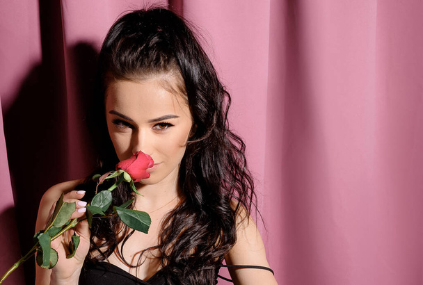 Young beautiful brunette girl in a black dress sniffs a red rose - Foto, Imagen
