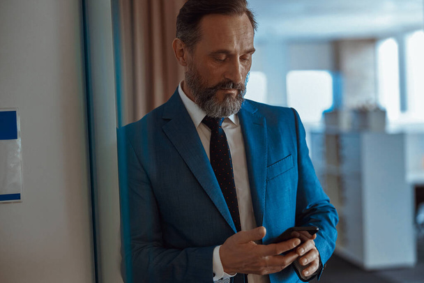 Bearded businessman reading message on phone in modern office - Foto, afbeelding