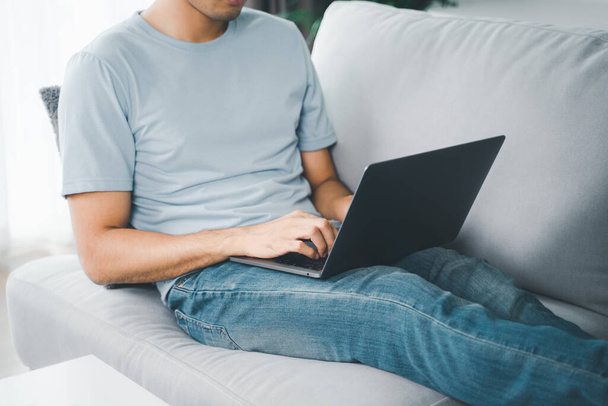 Młody człowiek siedzący na kanapie w domu piszący na klawiaturze laptopa do pracy online, korzystający z internetu, freelancer, zrelaksowany, nowoczesny styl życia pracy, praca w domu - Zdjęcie, obraz