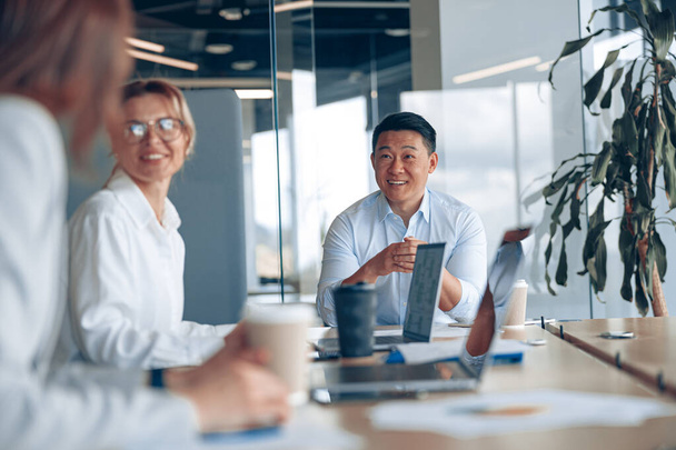 Молодой азиатский бизнесмен подслушивает своих коллег на встрече в современном офисе. Концепция командной работы - Фото, изображение