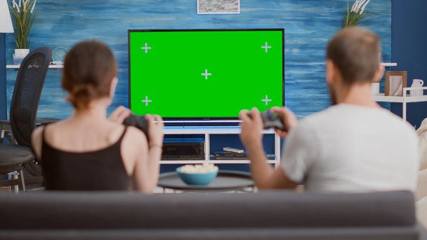 Pari tilalla ohjaimet pelaa konsolia online-pelejä vihreällä näytöllä tv istuu sohvalla modernissa olohuoneessa. Nuori nainen ja poikaystävä viettää vapaa-aikaa pelaamista chroma avain mockup näyttö. - Valokuva, kuva