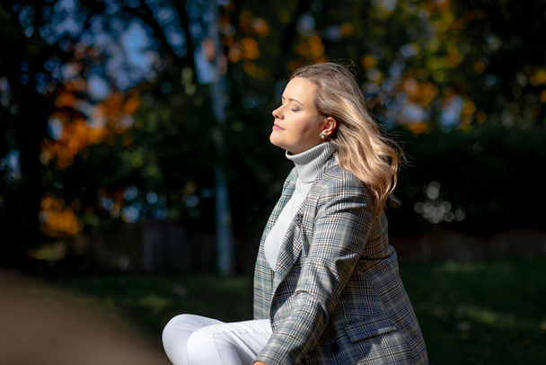 Портрет щасливої жінки, яка насолоджується сидячи в осінньому міському парку на сонячному дні вид збоку. Ділова леді в повсякденному одязі розслабляється і ходить на вулиці, на природі. Затишна золота осінь і погода
 - Фото, зображення