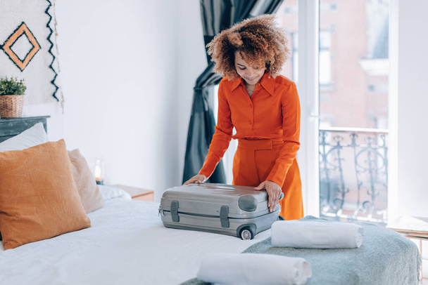 curly hair brazilian elegant young woman in orange suit open her luggage in hotel room bed - Fotografie, Obrázek