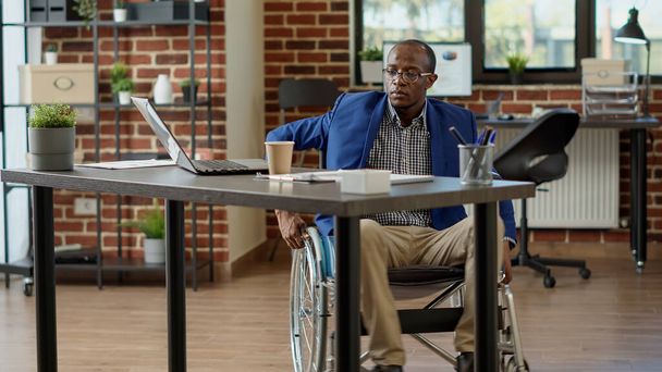 Gelähmter Arbeiter mit körperlicher Behinderung, der Statistiken am Laptop analysiert. Rollstuhlfahrer mit Beeinträchtigung arbeitet mit chronischer Krankheit, lebt mit Behinderung am Arbeitsplatz. - Foto, Bild