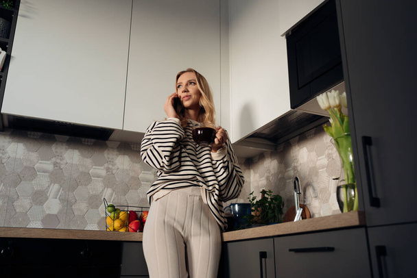 Young happy woman holding cup of coffee and smiling speaking by smart phone. Portrait from below of lady in home clothes having breakfast in modern design kitchen. Dieting and proper nutrition - Foto, Imagen