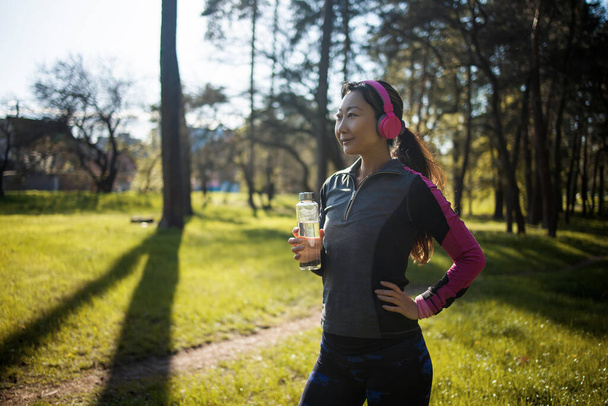 Fiatal ázsiai nő sportruházatban ivóvíz egy üvegből, miközben kocog a városi parkban reggel. Egészséges női sportoló élvezni szabadtéri életmód sport edzés edzés futás gyakorlat - Fotó, kép