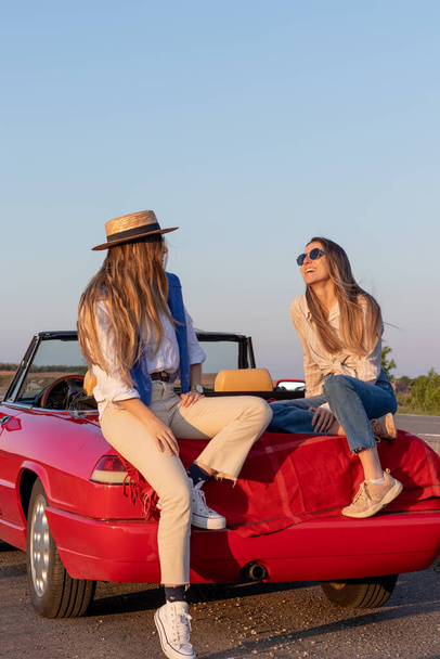 Heureux amis féminins acclamant assis dans un carat rouge convertible coucher de soleil avec lumière dorée et fusée éclairante avec espace de copie - Photo, image