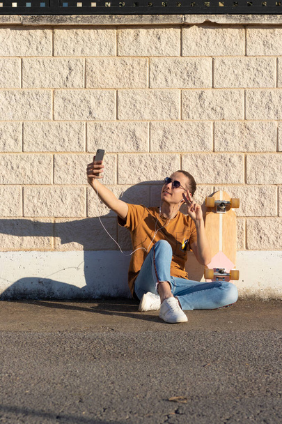 Joven tomando una foto selfie con orgulloso de su monopatín para enviar a sus amigos mientras está sentado en el suelo en la calle - Foto, imagen