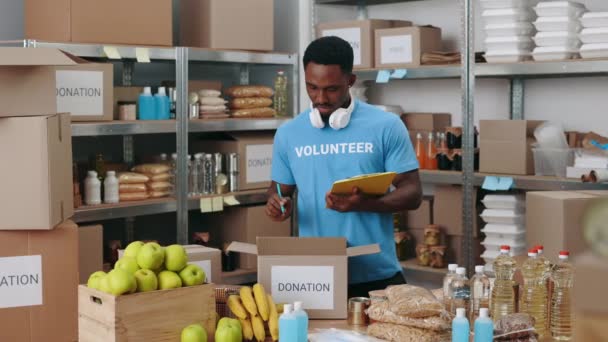 African american man in blue volunteer t-shirt using clipboard for taking notes while packing donation boxes at storage. Concept of people and volunteering. - Кадры, видео