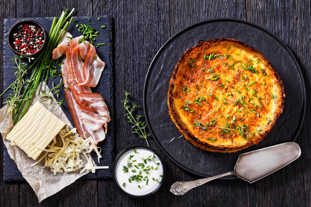 Hash Brown crust bacon and cheddar quiche on black plate with chives, sliced bacon, thyme, piece of cheddar cheese on stone board on dark oak table, horizontal view from above, flat lay - 写真・画像