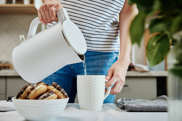 Время чая, женщина наливает кипяченую воду из электрического чайника в чашку для приготовления чая, Утренний завтрак на кухне - Фото, изображение