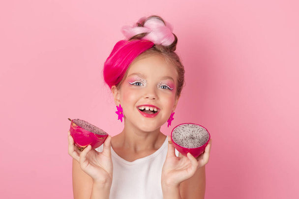 Κοντινό πλάνο πορτρέτο του όμορφου κοριτσιού με ροζ χτένισμα με φρούτα δράκο σε ροζ φόντο. Στούντιο πλάνο του γοητευτικό κορίτσι μεταξύ με ροζ μακιγιάζ απολαμβάνοντας ζουμερό κόκκινο pitaya. εξωτικά φρούτα Pitahaya. - Φωτογραφία, εικόνα