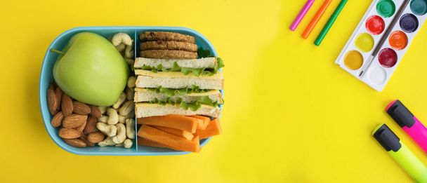 Een spandoek. Lunch in de blauwe doos en schoolbriefpapier op de gele achtergrond. Bovenaanzicht. Kopieerruimte. - Foto, afbeelding