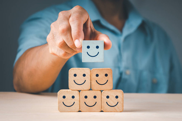 男の手は笑顔のシンボルを持つ木製の立方体ブロックフォームを配置します。満足度調査の概念は、顧客体験を評価する最高の企業サービスに基づいています. - 写真・画像