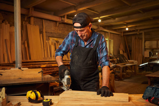 Ανώτερος Επαγγελματίας ξυλουργός σε ομοιόμορφο ξύλο πριονίσματος με ηλεκτρικό παζλ στο εργαστήριο ξυλουργικής - Φωτογραφία, εικόνα