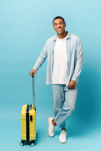 Full length photo of smiling young African-American guy in casual denim wear standing with yellow suitcase - Photo, Image