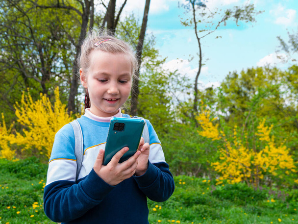 Adorable child girl chatting talking in social media smartphone learning language reading books outdoor. Blogger kid with gadget distance school online podcast lesson cell phone in park yellow flowers - Photo, Image