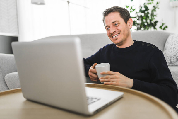 European man holding coffee while working on his laptop in bright living room. - Foto, imagen