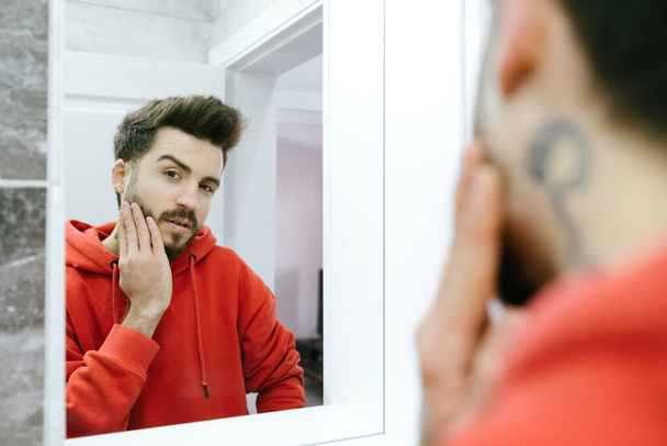 Attractive bearded young man looking to camera while touching face applying moisturizer on face, standing near mirror in modern bathroom. Male facial skincare routine. - Фото, изображение