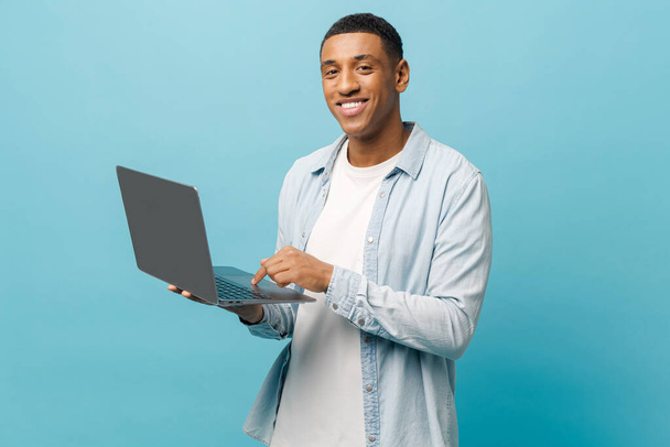 Optimistic african-american male student in casual jeans shirt using laptop pc isolated on blue - Valokuva, kuva
